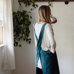 May include: A teal-colored apron with cross-back straps. The apron is made of a textured fabric and is worn over a white long-sleeved shirt and blue jeans. The apron is a kitchen or craft item.