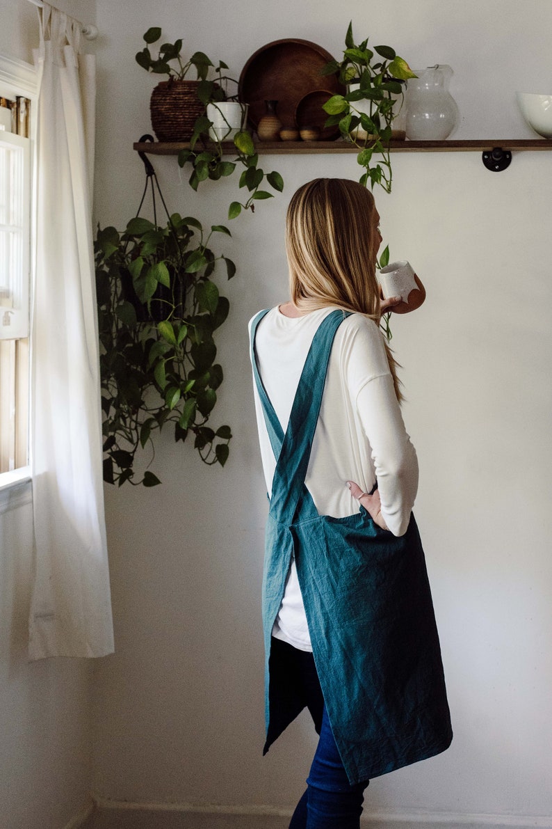 May include: A teal-colored apron with cross-back straps. The apron is made of a textured fabric and is worn over a white long-sleeved shirt and blue jeans. The apron is a kitchen or craft item.