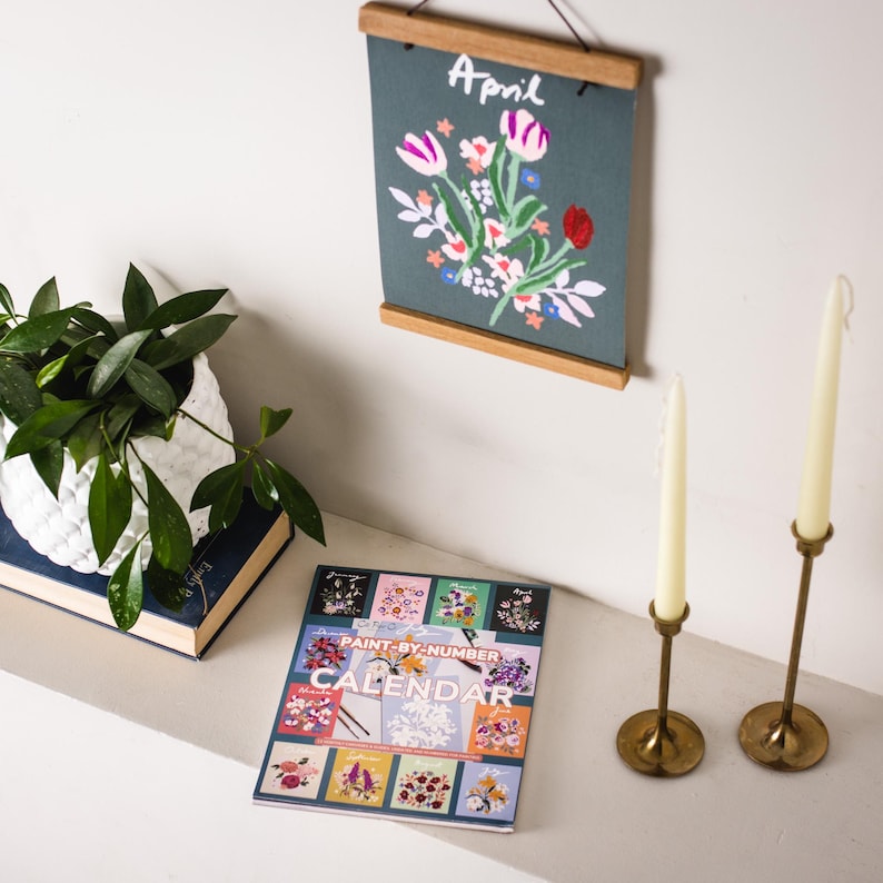 May include: A floral-themed home decor arrangement featuring a framed artwork with the word "April" and a flower design. A "Paint-by-Number" calendar is displayed, along with two brass candle holders with white candles and a potted plant.