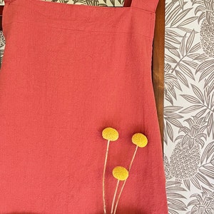 May include: A coral-colored apron with a large front pocket and shoulder straps. The apron is made of textured fabric and is adorned with three yellow billy button flowers. The background features a pineapple-patterned wallpaper.
