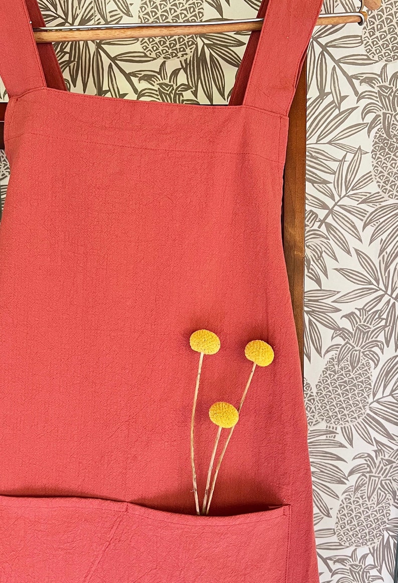 May include: A coral-colored apron with a large front pocket and shoulder straps. The apron is made of textured fabric and is adorned with three yellow billy button flowers. The background features a pineapple-patterned wallpaper.