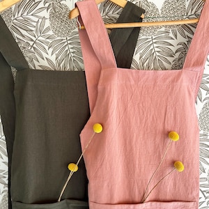 May include: Two aprons hanging on wooden hangers against a patterned background. One apron is a dusty rose color, and the other is a dark olive green. Each apron has a large front pocket and is decorated with yellow flowers.