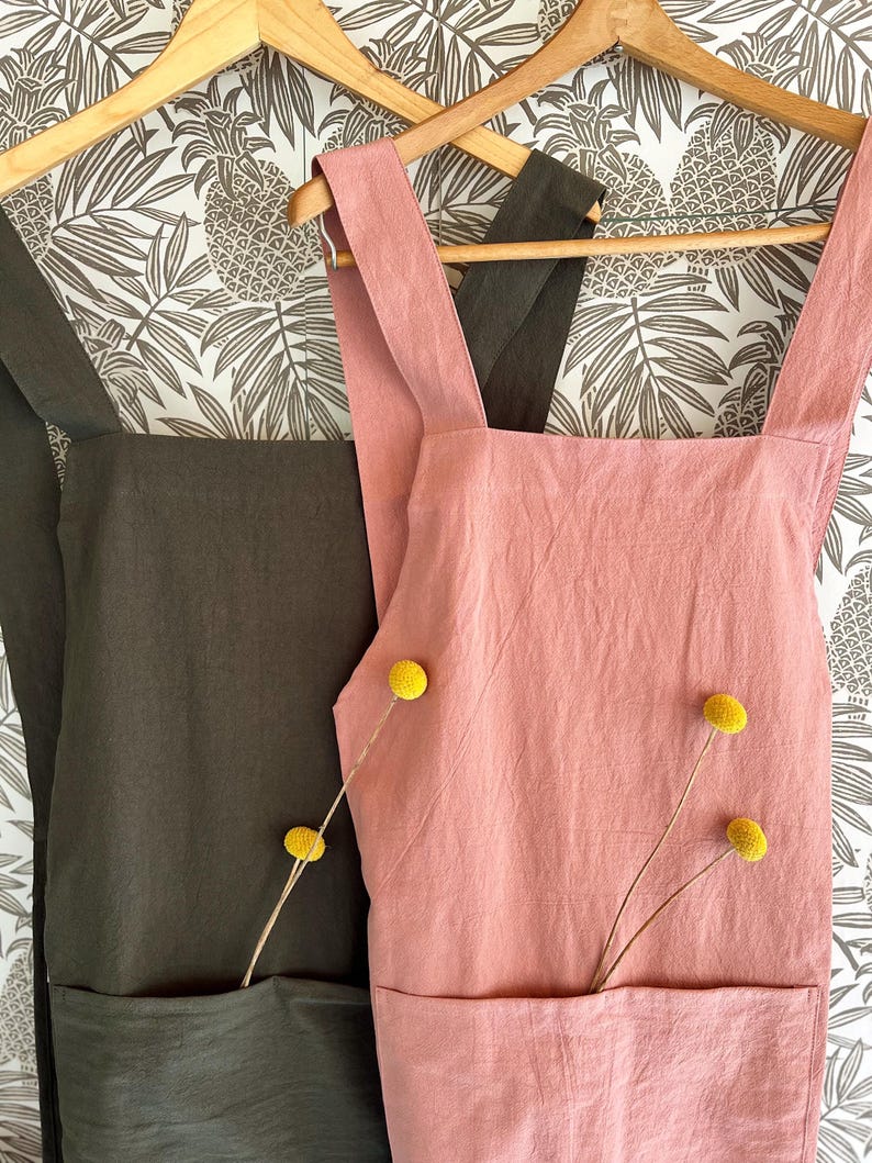 May include: Two aprons hanging on wooden hangers against a patterned background. One apron is a dusty rose color, and the other is a dark olive green. Each apron has a large front pocket and is decorated with yellow flowers.