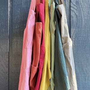 May include: A collection of colorful aprons hanging on a string against a blue wooden background. The aprons are in shades of pink, red, yellow, green, and blue. Each apron has a pocket.