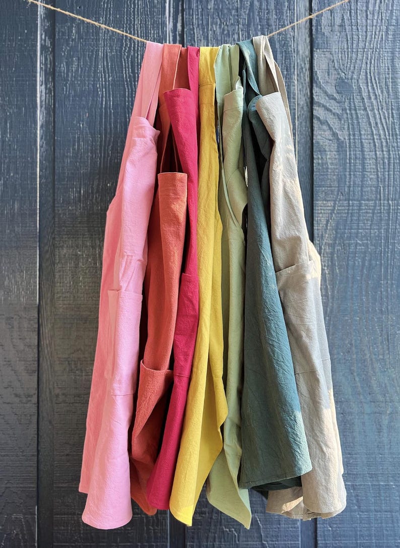 May include: A collection of colorful aprons hanging on a string against a blue wooden background. The aprons are in shades of pink, red, yellow, green, and blue. Each apron has a pocket.