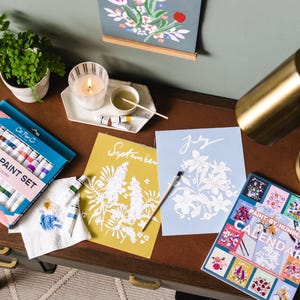 May include: A desk with art supplies, including an acrylic paint set, paint-by-number calendar, and floral art prints. A lit candle, small potted plant, and a brass desk lamp are also on the desk.