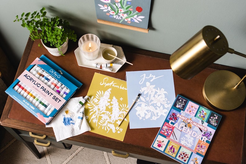 May include: A desk with art supplies, including an acrylic paint set, paint-by-number calendar, and floral art prints. A lit candle, small potted plant, and a brass desk lamp are also on the desk.