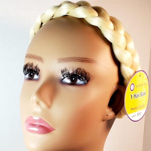 Puede incluir: Una diadema de trenza rubia sobre una cabeza de maniquí. La trenza sintética es de un tono platino claro. Lleva una etiqueta circular morada y amarilla con el texto Master Piece Y Hair Band Premium Quality color 613.
