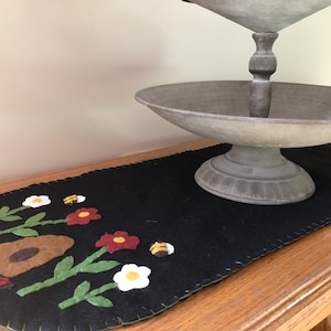 May include: A black felt table runner with a floral design featuring white and red flowers, green leaves, and brown and yellow bees. The runner is on a wooden table with a gray metal tiered serving tray.
