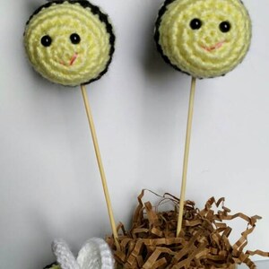May include: Three crocheted bumblebees with black and yellow stripes and white wings on wooden skewers. The bees are arranged in a glass jar filled with brown shredded paper.