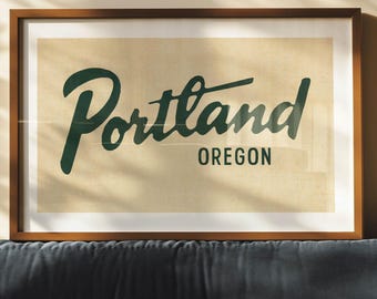 Puede incluir: Un cuadro enmarcado con la palabra "Portland" en verde oscuro, con "Oregon" debajo. El cuadro tiene un fondo beige y un marco de madera marrón oscuro. El cuadro se muestra sobre un sofá azul oscuro.