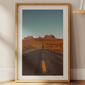 May include: A framed photograph of a long, straight road leading towards a mountain range in the distance. The road is paved and has a yellow line down the center. The mountains are red and brown and are silhouetted against a blue sky.