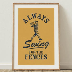 May include: Framed print with a mustard-yellow background and the text "ALWAYS Swing FOR THE FENCES" in navy blue. A baseball player is illustrated in the center. The print is in a wooden frame and leans against a white wall.
