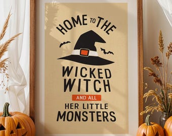 May include: A framed Halloween print with the text "Home to the Wicked Witch and All Her Little Monsters" is displayed. Two carved pumpkins and decorative dried grasses are in front of the print. The pumpkins are orange and have carved faces.