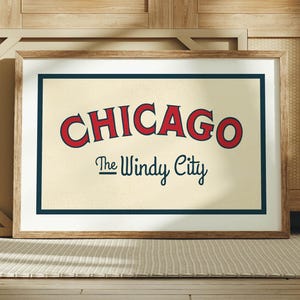 Op de afbeelding: Ingelijste kunstdruk met het woord "CHICAGO" in rode, gebogen letters boven "The Windy City" in een scriptlettertype. Het kunstwerk heeft een cr&egrave;mekleurige achtergrond met een marineblauwe rand en een houten lijst. De print wordt op een tapijt weergegeven.