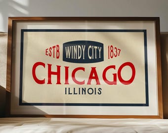 May include: Framed art print featuring the text "CHICAGO ILLINOIS" in red, with "ESTB WINDY CITY 1837" above. The artwork has a cream background and a dark blue border, set in a wooden frame. The print rests on a beige sofa.