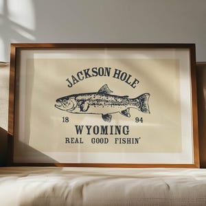 May include: Framed art print featuring a detailed illustration of a fish, with the text "JACKSON HOLE WYOMING REAL GOOD FISHIN'". The artwork is in a wooden frame and rests on a beige sofa. The print has a vintage aesthetic.