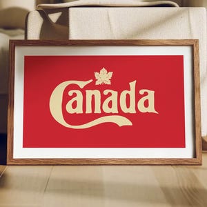 Puede incluir: Impresión enmarcada con la palabra "Canada" en letras color crema y una hoja de arce sobre un fondo rojo. La impresión está en un marco de madera, apoyada sobre un suelo de madera. Cerca de un sofá.