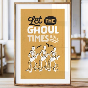 May include: Framed art print with a mustard yellow background featuring the text "Let the Ghoul Times Roll." Three skeleton figures play guitars below the text. The artwork is in a wooden frame and sits on a wooden table.
