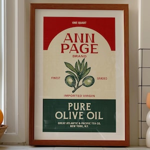 May include: A framed vintage-style print for "Ann Page Pure Olive Oil." The print features a red top banner, a central olive branch illustration, and text in a classic font. The print is displayed on a tiled countertop.