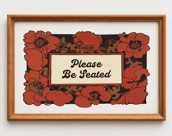 May include: A framed print with the text "Please Be Seated". The artwork features a rectangular cream-colored center with the text, surrounded by a border of red flowers and dark brown foliage, all set against a black background.