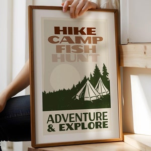 May include: A framed print with a vintage aesthetic featuring the words "HIKE CAMP FISH HUNT ADVENTURE & EXPLORE" in brown and green text. The print also features a green forest with two white tents and a white moon.
