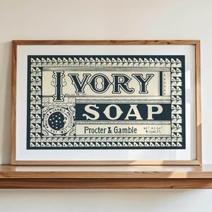 Puede incluir: Una impresión enmarcada de un anuncio de jabón Ivory. La impresión presenta las palabras "Ivory Soap" en letras grandes y en negrita, con texto adicional que indica la marca y el peso neto. El marco es de color marrón claro y la impresión está sobre una estantería de madera.