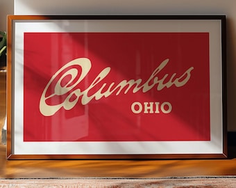 May include: Framed art print featuring the word "Columbus" in a cursive font, with "OHIO" below, set against a red background. The print is in a wooden frame and rests on a wooden floor.