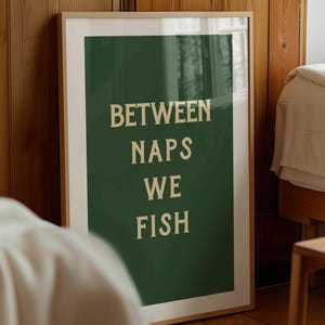 May include: A framed art print with the text "BETWEEN NAPS WE FISH" in a white, bold font on a dark green background. The print is in a light wood frame and leans against a wooden wall.