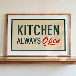 May include: A framed sign with the words "KITCHEN ALWAYS Open" in dark green and red lettering. The sign is in a wooden frame and sits on a wooden shelf against a white wall. The sign has a vintage aesthetic.