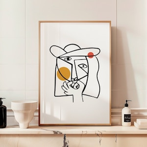 Puede incluir: Una impresión en blanco y negro de arte de línea de una cara estilizada con un sombrero, un círculo amarillo y un círculo rojo. La impresión está enmarcada en un marco de madera claro y está colgada en una pared de azulejos blancos.