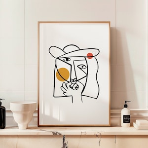 May include: A black and white line art print of a person's face with a hat, a yellow circle, and a red circle. The print is framed in a light wood frame and is hanging on a white tiled wall.