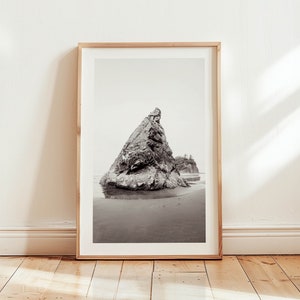 May include: A black and white photograph of a large rock formation on a beach. The rock is tall and pointed, and the beach is sandy. The photo is framed in a light wood frame.