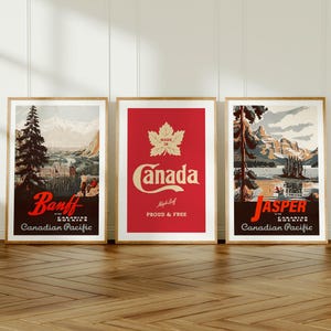 Op de afbeelding: Drie ingelijste vintage reisposters. De posters tonen schilderachtige uitzichten op Banff en Jasper, Canada, en een rode poster met het woord "Canada" en een esdoornblad. De posters zitten in houten frames en worden op een houten vloer getoond.