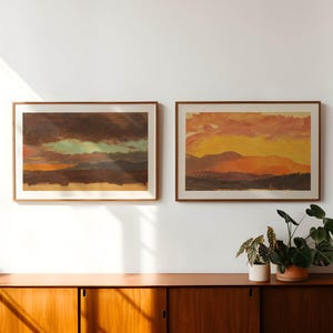 May include: Two framed landscape paintings with sunset scenes. One features dark clouds and a hint of green, the other a vibrant orange and yellow sky. The paintings hang above a wooden cabinet.