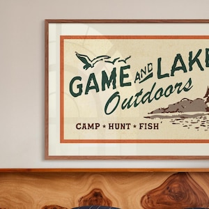 Op de afbeelding: Een vintage poster met een bruine houten lijst. De poster heeft een beige achtergrond met een rode rand en groene tekst die "GAME AND LAKE Outdoors CAMP * HUNT * FISH" luidt. De poster toont ook een tekening van een meer met een berg op de achtergrond en twee vogels die erboven vliegen.