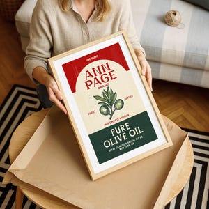May include: A framed print featuring a vintage-style olive oil label. The label, in red, beige, and green, reads "ANN PAGE BRAND" and "PURE OLIVE OIL." The print is held by a person, and the frame is light wood.