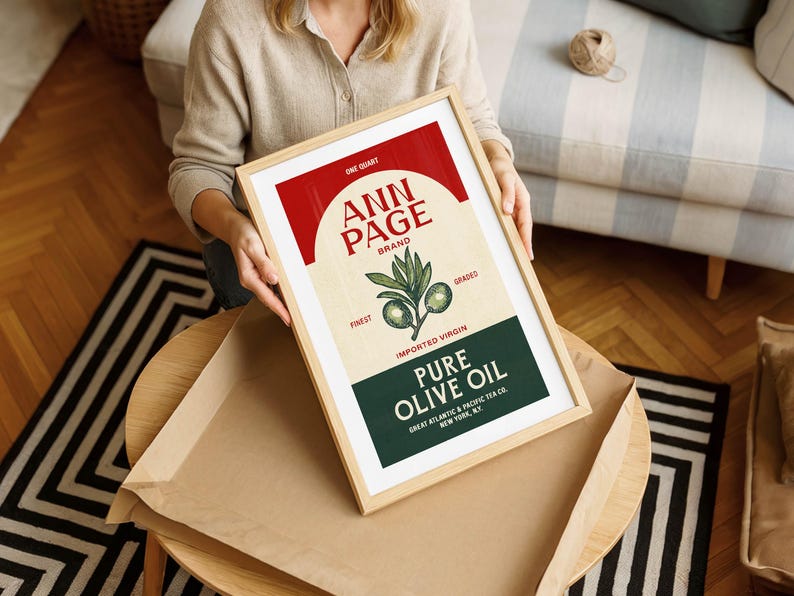 May include: A framed print featuring a vintage-style olive oil label. The label, in red, beige, and green, reads "ANN PAGE BRAND" and "PURE OLIVE OIL." The print is held by a person, and the frame is light wood.