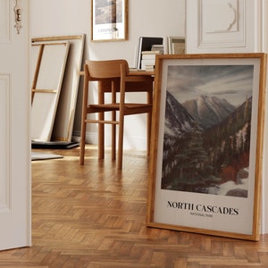 K&ouml;nnte beinhalten: Ein gerahmtes Poster mit einem Foto einer Bergkette mit schneebedeckten Gipfeln und einem Wald im Vordergrund. Das Poster tr&auml;gt den Titel "North Cascades National Park".