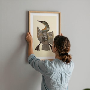May include: A framed art print featuring a stylized illustration of a bird with outstretched wings. The artwork has a beige background and a brown wooden frame. The bird's plumage is detailed with shades of brown, gray, and blue. The print is being held against a gray wall.