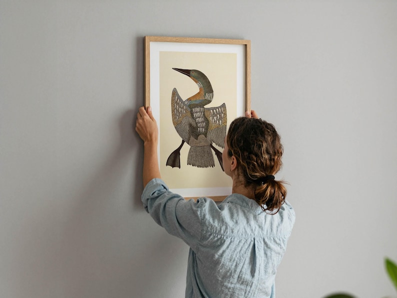 May include: A framed art print featuring a stylized illustration of a bird with outstretched wings. The artwork has a beige background and a brown wooden frame. The bird's plumage is detailed with shades of brown, gray, and blue. The print is being held against a gray wall.