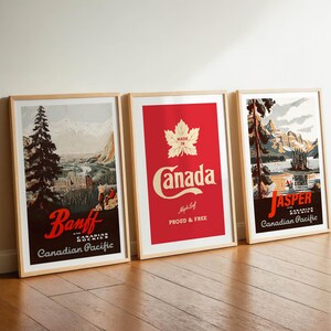 Op de afbeelding: Drie ingelijste reisposters leunen tegen een witte muur. De posters tonen vintage ontwerpen met de teksten "Banff", "Canada" en "Jasper". De "Canada" poster heeft een rode achtergrond met een esdoornblad.