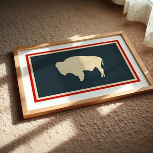 May include: A rectangular framed print featuring a stylized cream-colored silhouette of a bison centered on a dark blue background. The design is enclosed by a thin cream border and a bold red outer frame, all encased in a light wood frame.