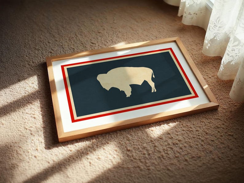 May include: A rectangular framed print featuring a stylized cream-colored silhouette of a bison centered on a dark blue background. The design is enclosed by a thin cream border and a bold red outer frame, all encased in a light wood frame.