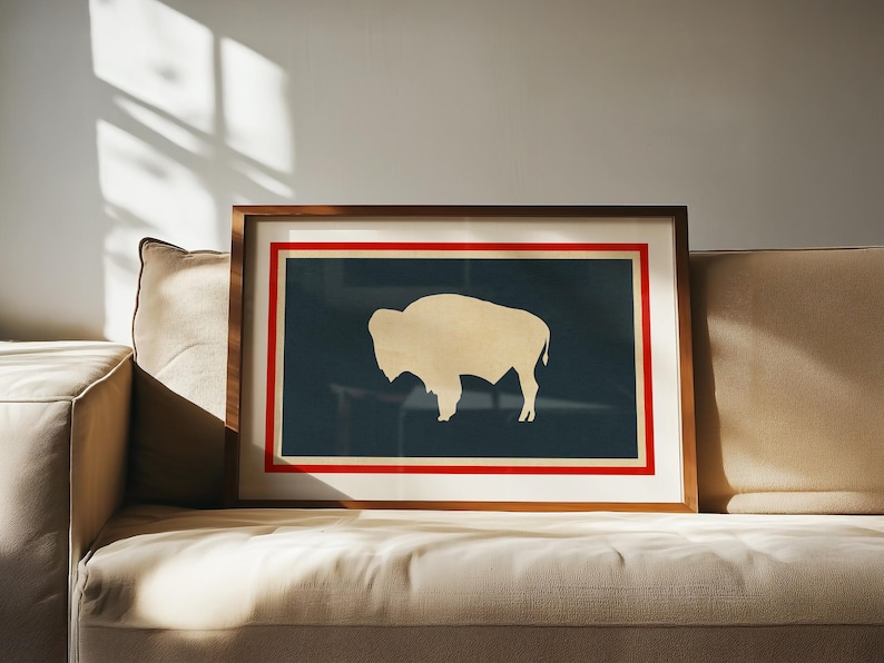May include: A framed rectangular wall art piece featuring a dark blue background with a cream-colored silhouette of a bison in the center. The artwork is enclosed by a thin red border and a white mat, all encased in a medium-toned wooden frame.