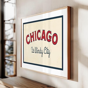Op de afbeelding: Een ingelijste print met het woord "CHICAGO" in rode, gebogen letters boven "The Windy City" in een scriptlettertype. Het kunstwerk heeft een cr&egrave;mekleurige achtergrond met een marineblauwe rand en een houten lijst.