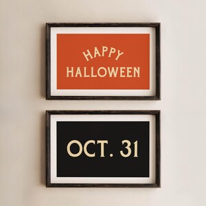 May include: Two framed Halloween-themed prints. The top print has an orange background with "HAPPY HALLOWEEN" in cream-colored text. The bottom print has a black background with "OCT. 31" in cream-colored text. Both are in dark brown frames.
