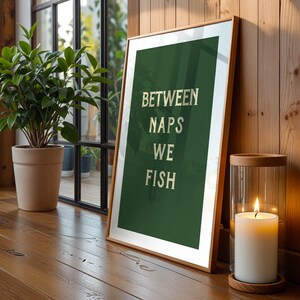 May include: A framed art print with the text "BETWEEN NAPS WE FISH" in white letters on a dark green background. The print is leaning against a wooden wall, next to a lit candle in a glass holder. A potted plant is in the background.
