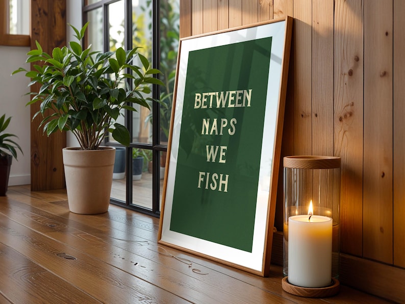 May include: A framed art print with the text "BETWEEN NAPS WE FISH" in white letters on a dark green background. The print is leaning against a wooden wall, next to a lit candle in a glass holder. A potted plant is in the background.
