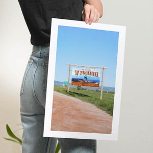 May include: A blue sky with a sign that reads "Welcome to Wyoming!" with a cowboy riding a horse on the sign. The sign is on a wooden post and is located on a dirt road with green grass on either side.